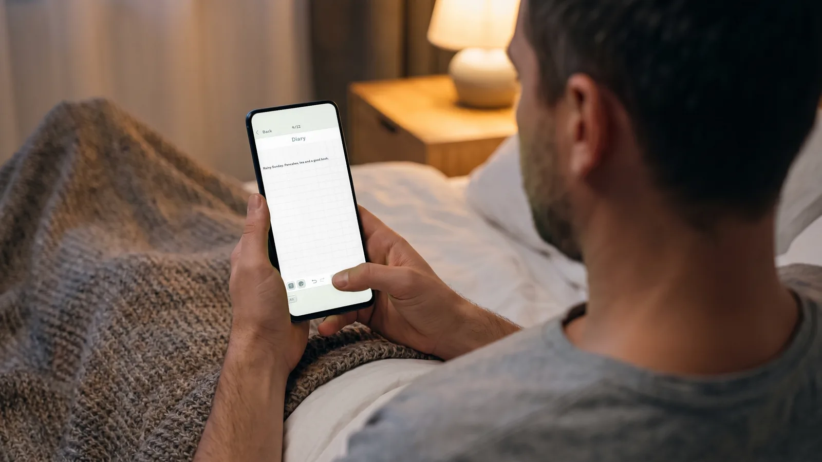 A man sitting up in bed in the evening, writing a short note on a DayBloq Diary page on his phone